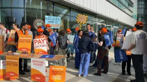 Dozens of people stand outside a hospital building holding placards and signs that say 'official picket' and wearing orange hats.