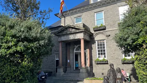 The Guernsey Information Centre. A grey building with coumns in the entrance with a statue of three bronze people in the bottom right. 