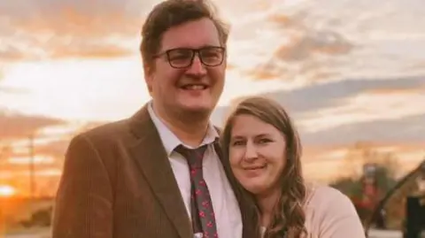 PA Media Edward and Laura Burr are smiling while standing close together outside. A sunset can be seen in the background. Edward has short brown hair and glasses and Laura has long brown hair.