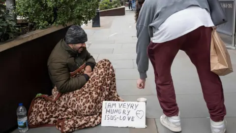 Generic image of a homeless person begging in the streets of London. Sign by his feet reads "I am very hungry"