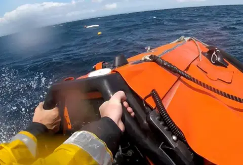 RNLI A woman in the distance holding onto a paddleboard at sea. The hands of a lifeboat crew member are visible in the foreground steering the boat 