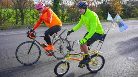 Paddy McGuiness cycling on a yellow 'chopper' bicycle on a tree-lined road on a sunny day. He is wearing a gree high-vis jacket and helmet and is joined by another cyclist wearing orange jacket.