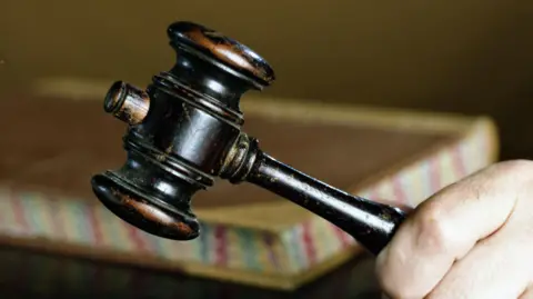 A person holds a gavel during an auction. A book can be seen resting on a table behind it.