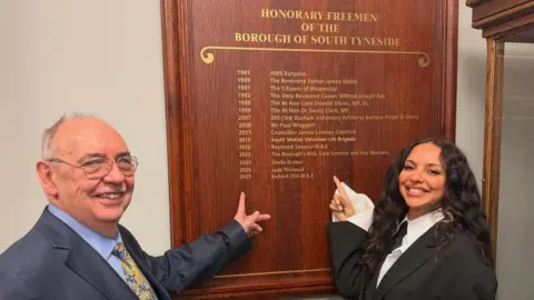 Richard Ord and Jade Thirlwall stand in front of a wooden plaque reading 'Honorary Freemen of the borough of South Tyneside'. Their names are inscribed on the plaque. The pair are pointing at the plaque and smiling. 