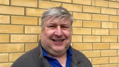 A man smiling, standing against a yellow brick wall. He is in a blue polo shirt and grey fleece and has short, grey hair.