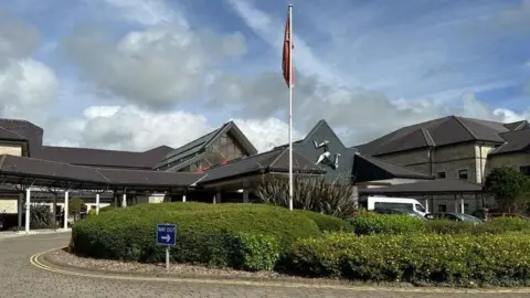 The front entrance to Noble's, which has a flag pole and greenery in front of it and a three legs of Man symbol on the roof.