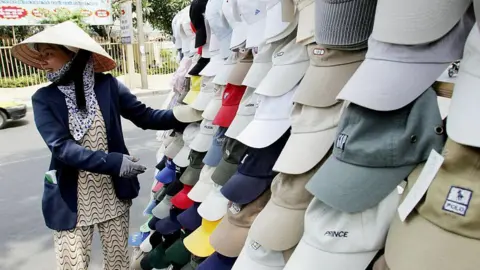 Getty Images Caps bearing the logos of Polo, GAP, Prince and others hang in the foreground next to a woman wearing a blue blazer and a straw nón lá hat
