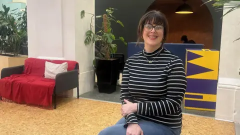 Mags White-O'Kane is sitting down in a colourful space. She is wearing a blue and white striped jumper and blue jeans. She has dark hair and is wearing glasses. In the background there is a double seat with a red throw over it and a plant on the right hand side. 