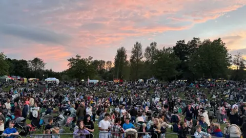 A crowd of people gathered in a field as the sun is setting. People are standing and sitting. The grass is almost completely covered due to the amount of people. There is a line of trees in the background and the sun is setting so the sky is pale blue with pink clouds.