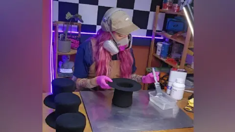 Hojo Hattery/Holly Johnson A woman with bright pink hair and a grey baseball cap on her head, is working on the bottom of a small black top hat. She has an air filter mask across her nose and mouth while her left hand holds a paint brush which she is dipping into a box containing clear liquid. Several small black top hats are sat next to her on the table.