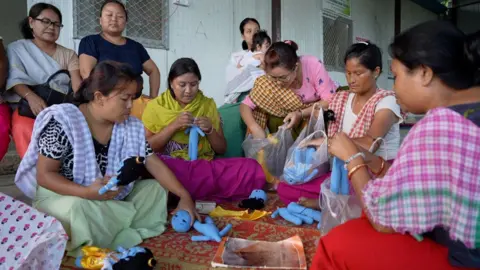 Midhat Ullah Hasani Woman packing blue crochet dolls. Four of them are sitting on the floor while others are sitting on tables and chairs.