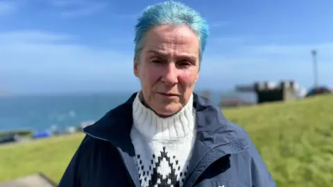 Sue Biggerstad has short bright blue hair, and is wearing a white woolly jumper under a navy jacket. She's standing in front of a green field with the sea in the background on a sunny day.