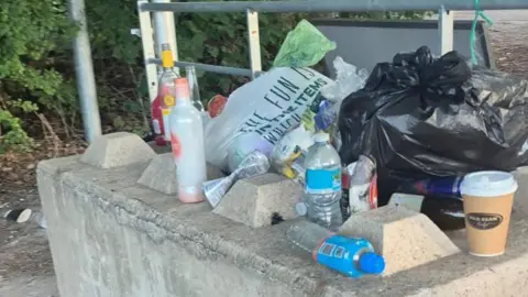 Rubbish bags, used plastic bottles and glass bottles and coffee cups, are left on a concrete barrier on a pathway. 