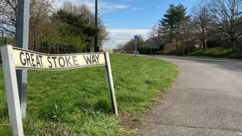 A roadsign saying Great Stoke Way, next to a grass verge on the left and a road on the right. The road sign is bent from an impact.