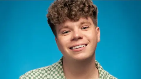 Daniel Swift with brown curly hair and wearing a green and white checked shirt stands in front of a blue background