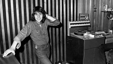 Mark Wesley Black and white image of Mark in the 70s in a recording studio. He has shoulder-length hair and is smiling at the camera.