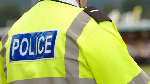 The back of a police officer who is wearing a high-viz yellow jacket with grey lining. The word POLICE is spelled out in white letters on a blue background on a panel towards the top of his back