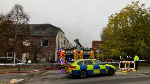 Firefighters and police officers work behind a cordon with police tape blocking off the area where two fire engines are situated. Several police officers can be seen to the right next to a large tree.
