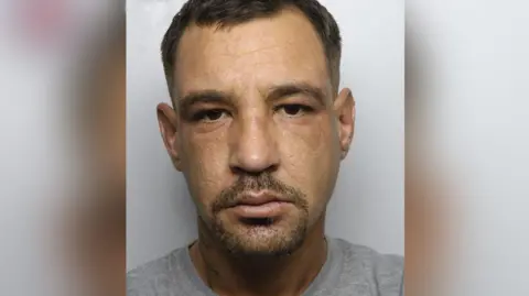 Police custody image of Martin Davies who has short dark brown hair and beard and is wearing a grey prison T-shirt.