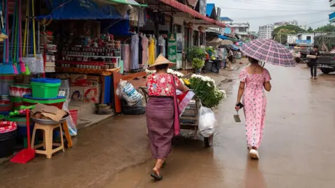 Jonathan Head/BBC A market in Shwe Kokko. Two women walk along a wet brown road, one pushing a wooden cart with white flowers in it. Their outfits are shades of pink. Shops selling baskets and dresses line the street on one side