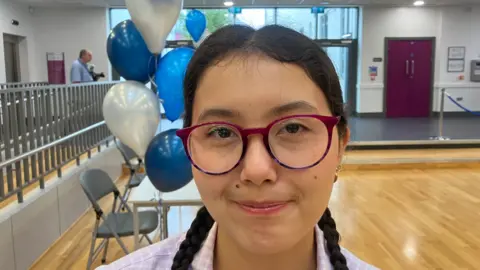 A girl, Anna McCauseland pictured in her school.
She is wearing purple glasses and has brunette hair in two plaits.