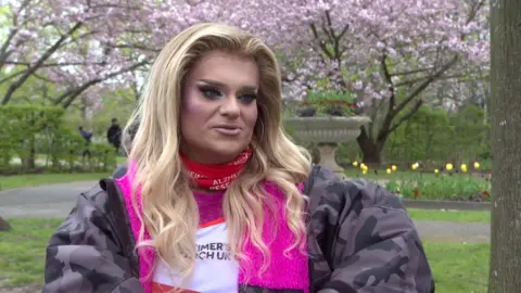 Drag Queen Kitty, with a blond wig, sits in the park wearing a pink dry robe. 