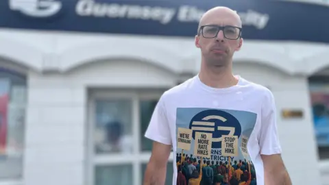 A bald man wearing glasses stands in front of Guernsey Electricity's head office. He is wearing a T-shirt with placards saying: "We're struggling! No more rate hikes, Stop the Hike and Power to the People."