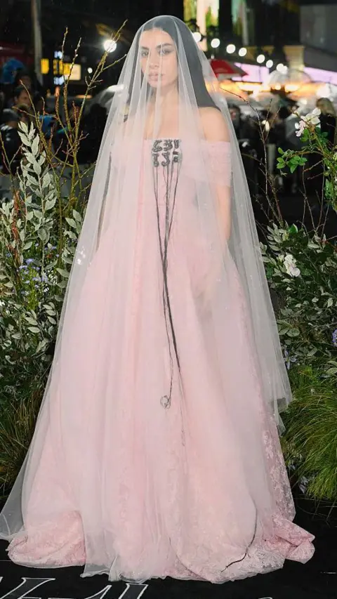 WireImage/Getty Images Charli XCX stands posing for a picture on the red carpet wearing a light pink floor-length dress with matching sheer veil that reaches the floor.
