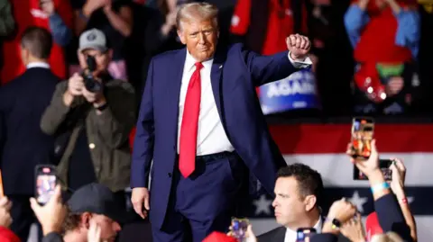 Getty Images Donald Trump walks through an arena crowd with his right fist raised out. Several mobile phones are photographing and videoing him.  