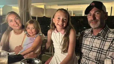 A family of four with two daughters pictured at a table during a meal. All are smiling.