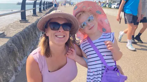 Marta and Ella on the promenade in Porthcawl. Martha wears a lavender vest, sunglasses and a hat and is crouched down next to Ella who wears a blue and white striped dress, pink hat and sunglasses