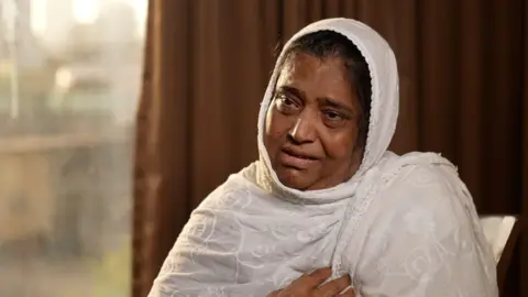 A womab wearing a white embroidered headscarf looks away from the camera. She is emotional and her eyes are teary. 