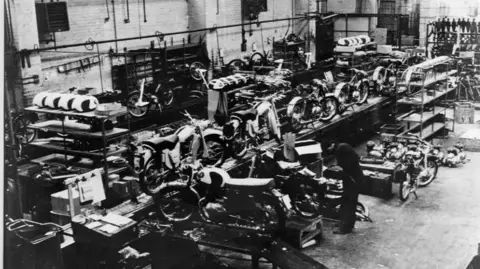 A production line of motorbikes, with several bikes and components sitting around. A man is tinkering with one of the vehicles in this black and white image, taken from above.