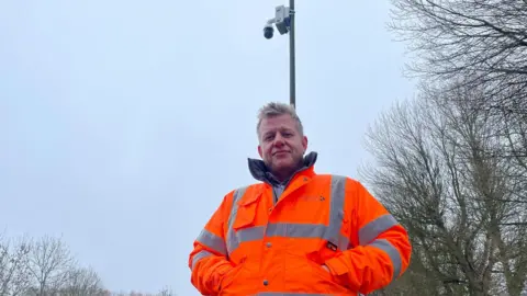 A man in a bright orange jacket stands below a lamp post with a camera attached to the top
