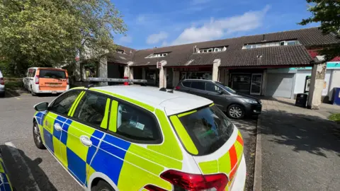 Stuart Woodward/BBC A police car parked outside a parade of shops