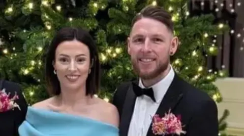 Kirsty Haywood A woman with dark hair and a blue dress and a man with ginger hair and a dark suit with a black bow tie, standing on front of a Christmas tree