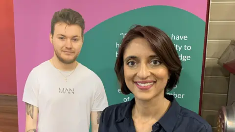 A woman stands in front of a poster inside a building. The woman has short dark brown hair and is wearing a dark blue shirt. The pink and green poster has a photo of a man wearing a white t-shirt on it.