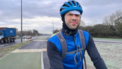 A biker is wearing a blue helmet and blue vest over cycling clothes. Behind him is a green cycle lane next to a road with cars, while on the other side a patch of green still has morning frost.