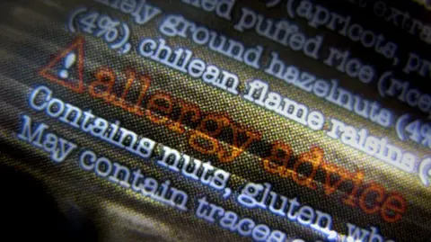 BBC The image shows a close-up of food packaging text, focusing on allergy information. The design includes a warning triangle with an exclamation mark, followed by the words “allergy advice” in bold orange-red lettering. Beneath that, the text states: “Contains nuts, gluten, wheat” and “May contain traces of seeds.”
