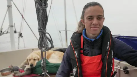 BBC Jazz Turner wears a life jacket and red and black waterproof clothing. She sits on her boat with ropes and a yellow Labrador in the background. She is looking at the camera smiling.