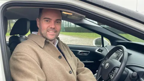 Dr Charlie Creissen sitting in the car's driving seat facing the camera. He's wearing a light brown coat.