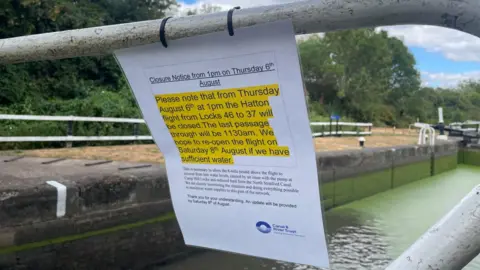 In the foreground is a paper sign cable tied to the metal railings of the lock with a closure notice from the Canal and River Trust on it. some of the text has been highlighted in yellow. Behind it is the closed lock.