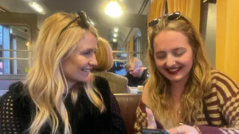 Family handout Judy Farnfield, who has blonde hair and sunglasses on her head, sat in a booth seat and looking to the side at her daughter Olivia Legg. Olivia is looking at her phone and smiling.