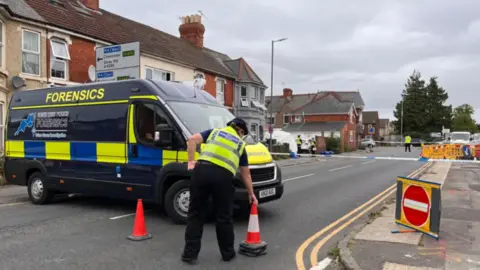 A police van with the word "forensics" on it. A police officer is laying out cones nearby.
