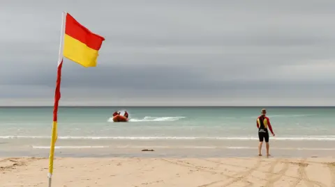A beach showing a flag marking the safe swimming area. A lifeguard is stood on the beach while looking at a rescue boat heading towards shore. 