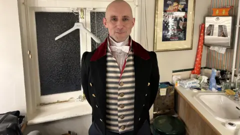 Jamie Tyler Jamie wearing a striped waistcoat with a jacket over the top and a white shirt and bowtie. He is in a dressing room with a sink on the right and a window behind him. He is smiling at the camera and has his hands behind his back.