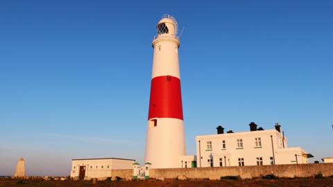 SATURDAY - Sunrise at Portland Bill lighthouse, the white and red light house building is glowing in the early morning sunshine. Behind there is a white building with a stone wall and to the left you can see the Trinity House Obelisk stone. There are no clouds in the clear blue sky overhead.