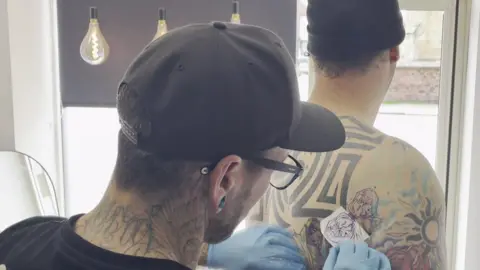 Tattoo artist Andy Walker preparing the artwork for a Marvel tattoo on Tom Radford's shoulder.