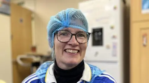 Shaun Whitmore/BBC A woman smiles at the camera. She has a blue hair net over her head and she is wearing glasses along with a blue shirt which has white stripes over the shoulders. 