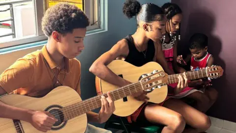 Cape Verdean children sit down and play guitars. Three are sat in a line.